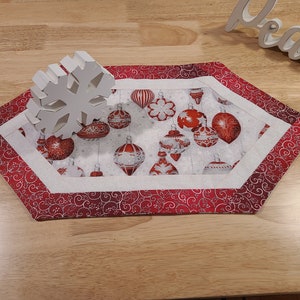 May include: A Christmas-themed table runner with a white snowflake decoration. The runner features a red and white ornament pattern on a white background, framed by a red border with a swirling design. The word "Peace" is visible in the background.