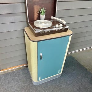 May include: A portable suitcase-style record player with a brown exterior and white platter sits atop a tall, rectangular cabinet. The cabinet features a light yellow frame, a teal-colored door with a silver handle, and a light wood-toned top surface.