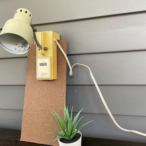 May include: A vintage yellow Mobilite lamp is mounted on a brown board, with a white cord. A small potted succulent sits on a dark wooden surface. The lamp has a metal shade and a yellow plastic base. The text "mobilite" is visible on the lamp.