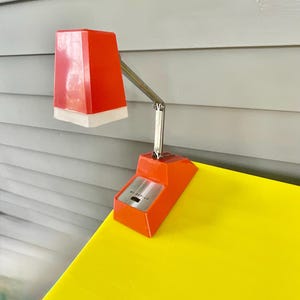 May include: A bright orange desk lamp with a rectangular shade and a white base trim. The lamp features a silver-toned adjustable metal arm and a rectangular orange base with a silver control panel labeled HI-OFF-LO for light intensity settings.