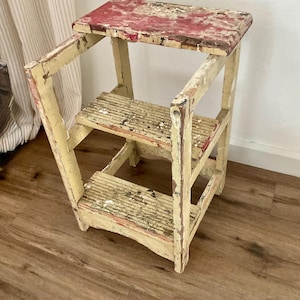 Puede incluir: Un taburete de madera de dos escalones con un aspecto envejecido. El escalón superior está pintado de rojo, mientras que el marco y los escalones son de color crema, con desgaste visible. El taburete está diseñado para alcanzar áreas elevadas.