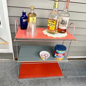 May include: A vintage, three-tiered serving cart with a pink top shelf, gray middle shelf, and red bottom shelf. The cart holds bottles of liquor, plastic cups, a bowl of candy, and a can of Planters peanuts.