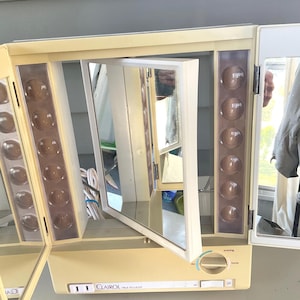 May include: A vintage Clairol True-to-Light tri-fold vanity mirror with a cream-colored plastic frame. The mirror features adjustable side panels, integrated light strips with circular bulbs, and a dial for light settings labeled day, office, home, and evening.