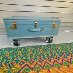 May include: A rectangular light blue suitcase with a flat top and rounded corners. It features three gold-toned metal latches, a matching light blue handle, and is mounted on four black swivel caster wheels for mobility and storage utility.