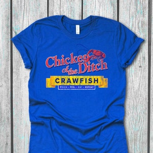 Puede incluir: Una camiseta azul real con un gráfico de estilo vintage que dice "Chicken of the Ditch Crawfish Pinch - Peel - Eat - Repeat". El gráfico presenta un cangrejo de río rojo y texto amarillo.