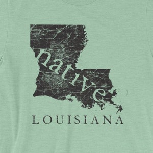 May include: A light green short-sleeve t-shirt featuring a black distressed graphic of the state of Louisiana. The word native is printed in a serif font across the map, and the word LOUISIANA is printed in capital letters below the map.