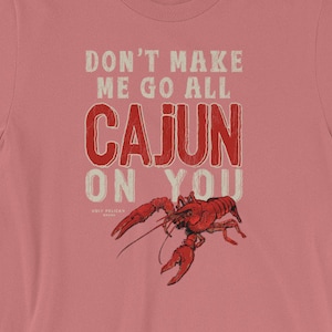 May include: A dusty rose short-sleeve t-shirt featuring a red crawfish graphic and the white and red text that reads DON'T MAKE ME GO ALL CAJUN ON YOU. The design is centered on the chest of the shirt, which has a crew neckline and short sleeves.
