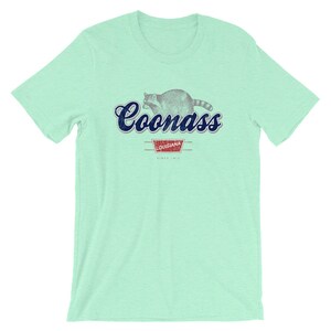 May include: A light green t-shirt with a blue and white graphic of a raccoon with the text "Coonass" and "Louisiana" in a red rectangle with the text "Since 1912".