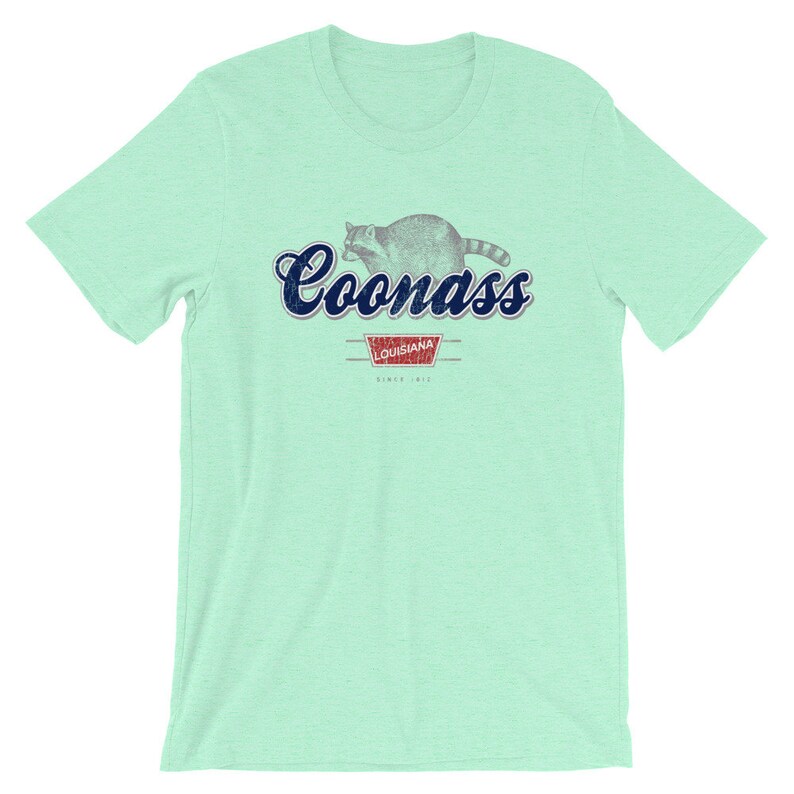 May include: A light green t-shirt with a blue and white graphic of a raccoon with the text "Coonass" and "Louisiana" in a red rectangle with the text "Since 1912".