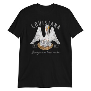 May include: A black short-sleeve t-shirt featuring a graphic of a white pelican in a nest with chicks. The text reads LOUISIANA, EST. 1812, and Laissez les bon temps rouler. The design uses white, grey, and orange colors with a distressed texture.