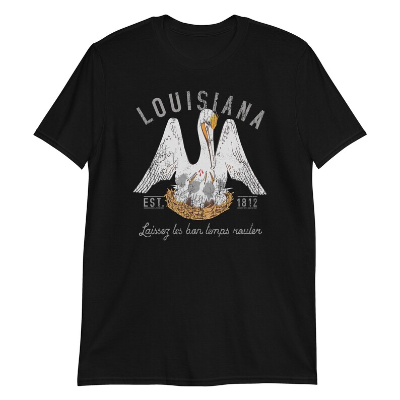 May include: A black short-sleeve t-shirt featuring a graphic of a white pelican in a nest with chicks. The text reads LOUISIANA, EST. 1812, and Laissez les bon temps rouler. The design uses white, grey, and orange colors with a distressed texture.