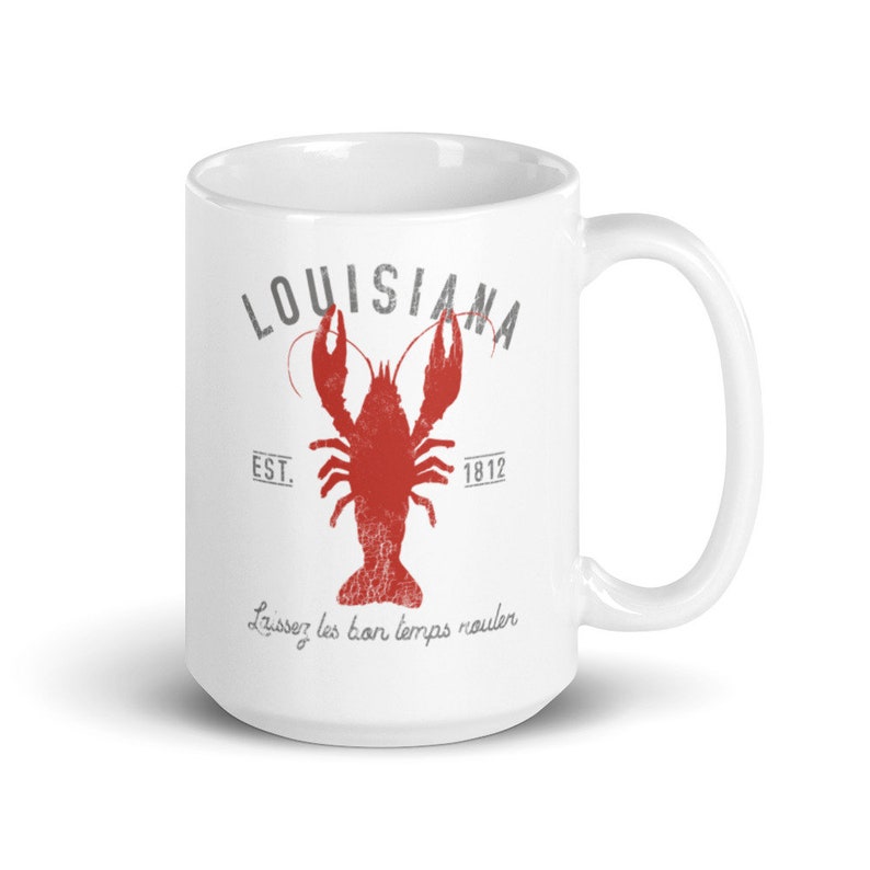 May include: A white ceramic mug featuring a red crawfish graphic in the center. Above the crawfish is the text LOUISIANA in gray, with EST. 1812 on either side. Below the crawfish, the text Laissez les bon temps rouler is printed in gray.