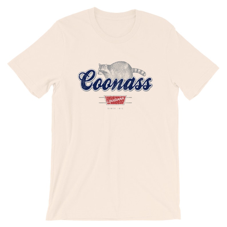 May include: A beige t-shirt with a blue and white graphic of a raccoon and the text "Coonass Louisiana Since 1912."