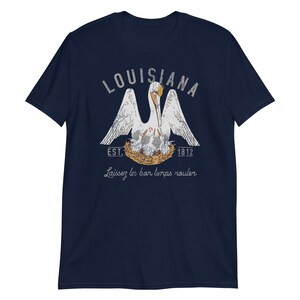 May include: A navy blue short-sleeve t-shirt featuring a graphic of a white pelican in a nest with chicks. The text reads LOUISIANA, EST. 1812, and Laissez les bon temps rouler. The design uses white, orange, and yellow colors with a distressed texture.