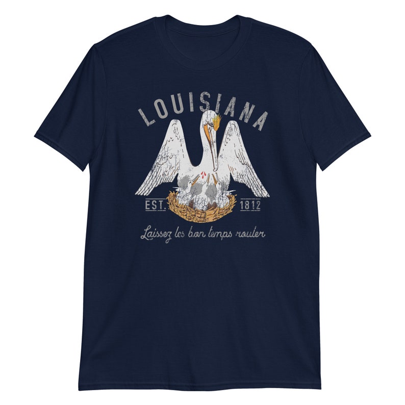 May include: A navy blue short-sleeve t-shirt featuring a graphic of a white pelican in a nest with chicks. The text reads LOUISIANA, EST. 1812, and Laissez les bon temps rouler. The design uses white, orange, and yellow colors with a distressed texture.