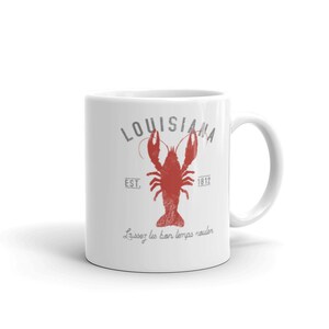 May include: A white ceramic mug featuring a red crawfish graphic in the center. Above the crawfish is the text "LOUISIANA" in grey, with "EST. 1812" on either side. Below the crawfish, the text "Laissez les bon temps rouler" is printed in grey.