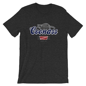 May include: A dark heather grey t-shirt with a blue and white graphic of a raccoon with the text "Coonass" and "Louisiana" in a vintage style.