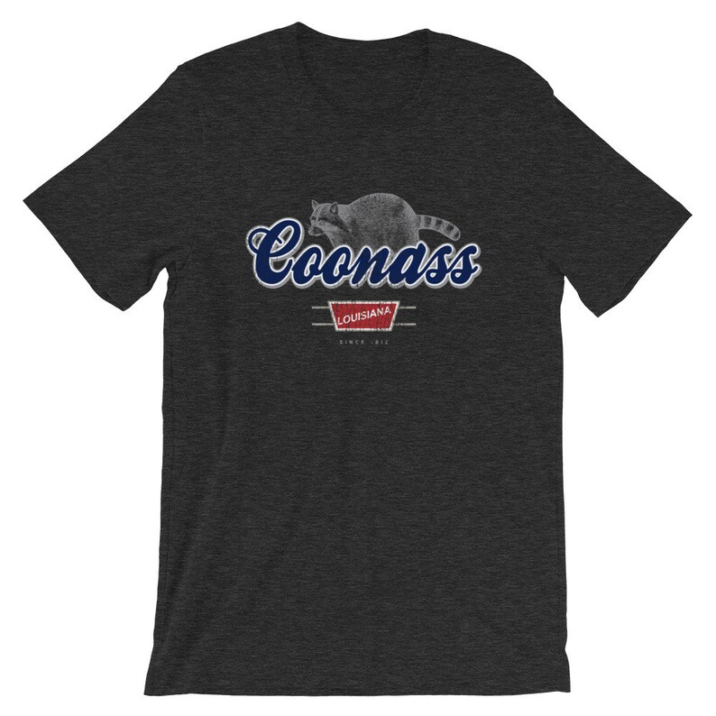 May include: A dark heather grey t-shirt with a blue and white graphic of a raccoon with the text "Coonass" and "Louisiana" in a vintage style.
