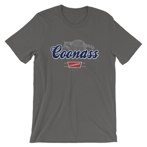 May include: A dark gray t-shirt with a blue and white graphic of a raccoon with the text "Coonass" in blue cursive lettering. The graphic also includes a red banner with the text "Louisiana" and "Since 1912".