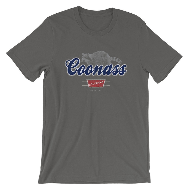 May include: A dark gray t-shirt with a blue and white graphic of a raccoon with the text "Coonass" in blue cursive lettering. The graphic also includes a red banner with the text "Louisiana" and "Since 1912".