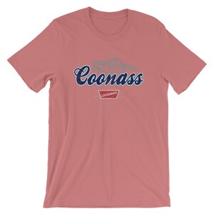 May include: A pink t-shirt with a blue graphic of a raccoon and the text "Coonass Louisiana".