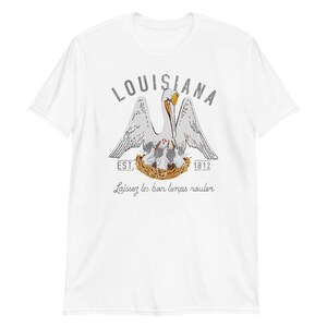 May include: A white short-sleeve t-shirt featuring a graphic of a pelican in a nest with chicks. The text reads LOUISIANA, EST. 1812, and Laissez les bon temps rouler. The pelican is white with orange accents, and the nest is brown.