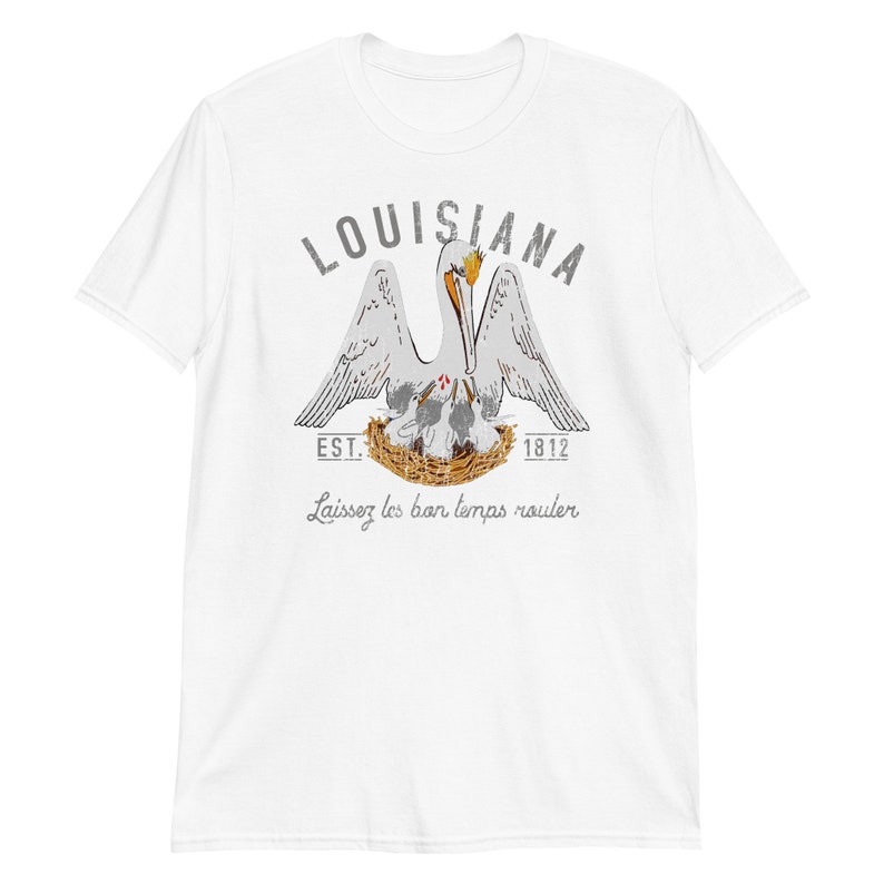 May include: A white short-sleeve t-shirt featuring a graphic of a pelican in a nest with chicks. The text reads LOUISIANA, EST. 1812, and Laissez les bon temps rouler. The pelican is white with orange accents, and the nest is brown.