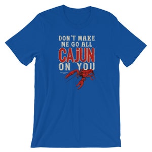 May include: A royal blue short-sleeve t-shirt featuring the white and red text DON'T MAKE ME GO ALL CAJUN ON YOU above a detailed illustration of a red crawfish. The design includes small text reading UGLY PELICAN BRAND below the crawfish.