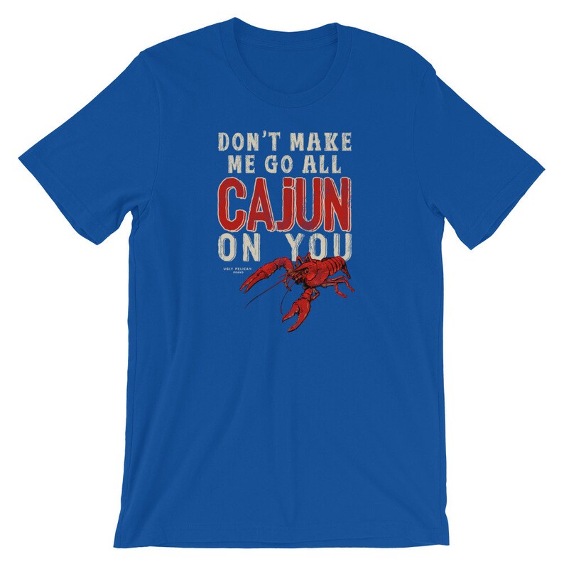 May include: A royal blue short-sleeve t-shirt featuring the white and red text DON'T MAKE ME GO ALL CAJUN ON YOU above a detailed illustration of a red crawfish. The design includes small text reading UGLY PELICAN BRAND below the crawfish.