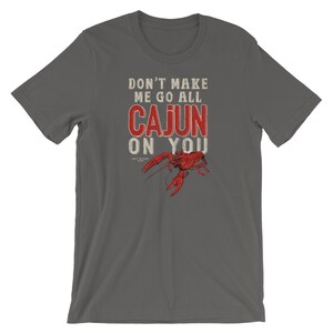 May include: A dark gray short-sleeve t-shirt featuring the white and red text "DON'T MAKE ME GO ALL CAJUN ON YOU" printed above a detailed illustration of a bright red crawfish. The design is centered on the chest of the garment.