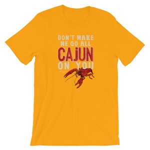 May include: A mustard yellow short-sleeve t-shirt featuring the white and red text DON'T MAKE ME GO ALL CAJUN ON YOU with a detailed illustration of a red crawfish positioned below the word CAJUN, centered on the front of the garment.