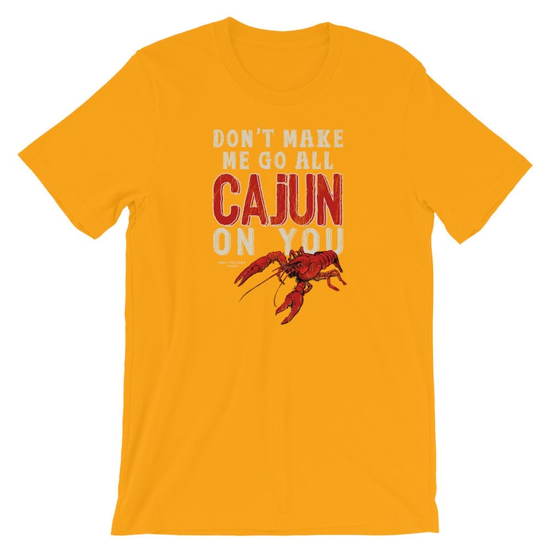 May include: A mustard yellow short-sleeve t-shirt featuring the white and red text DON'T MAKE ME GO ALL CAJUN ON YOU with a detailed illustration of a red crawfish positioned below the word CAJUN, centered on the front of the garment.