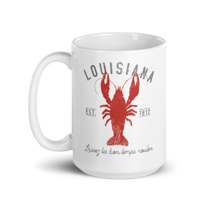 May include: A white ceramic mug featuring a red crawfish graphic in the center. Above the crawfish is the text LOUISIANA in grey, with EST. 1812 on either side. Below the crawfish is the text Laissez les bon temps rouler in grey script.