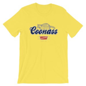 May include: Yellow t-shirt with a blue and white graphic of a raccoon with the text "Coonass" and "Louisiana" in a red rectangle.