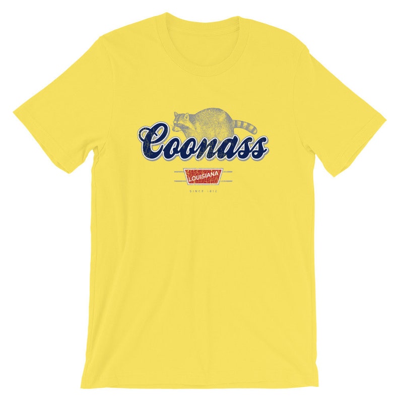 May include: Yellow t-shirt with a blue and white graphic of a raccoon with the text "Coonass" and "Louisiana" in a red rectangle.