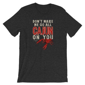 May include: A dark charcoal gray short-sleeve t-shirt featuring the white and red text DON'T MAKE ME GO ALL CAJUN ON YOU with a graphic of a red crawfish below the text. The design has a distressed, weathered appearance on the fabric.