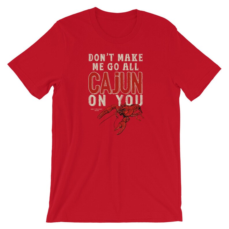 May include: A red short-sleeved t-shirt featuring the white text DON'T MAKE ME GO ALL CAJUN ON YOU and a detailed black illustration of a crawfish. Below the text, the small white text reads UGLY PELICAN BRAND, centered on the chest area.