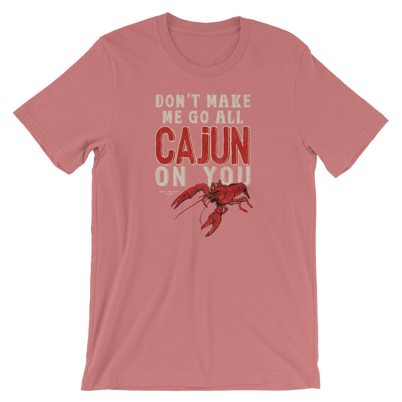 May include: A dusty rose short-sleeve t-shirt featuring a red crawfish graphic and the white and red text that reads DON'T MAKE ME GO ALL CAJUN ON YOU. The design is centered on the chest of the shirt, which has a crew neckline and short sleeves.