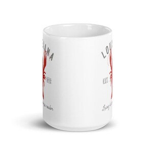 May include: A white ceramic mug featuring a red crawfish graphic, the text LOUISIANA, the year 1812, and the phrase Laissez les bons temps rouler in grey lettering. The design is printed on the side of the cylindrical mug with a handle.