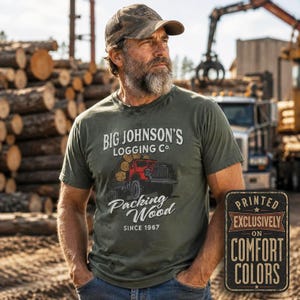 Pode incluir: Uma t-shirt verde-azeitona com um gráfico de um camião madeireiro vermelho a transportar troncos e o texto "BIG JOHNSON'S LOGGING CO. Packing Wood SINCE 1967". Um sinal retangular separado diz "PRINTED EXCLUSIVELY ON COMFORT COLORS".
