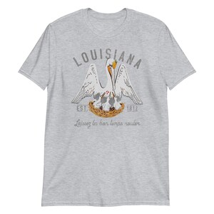 May include: A heather gray short-sleeve t-shirt featuring a graphic of a pelican in a nest with chicks. The text reads LOUISIANA, EST. 1812, and Laissez les bon temps rouler. The pelican is white with orange accents, and the nest is brown.