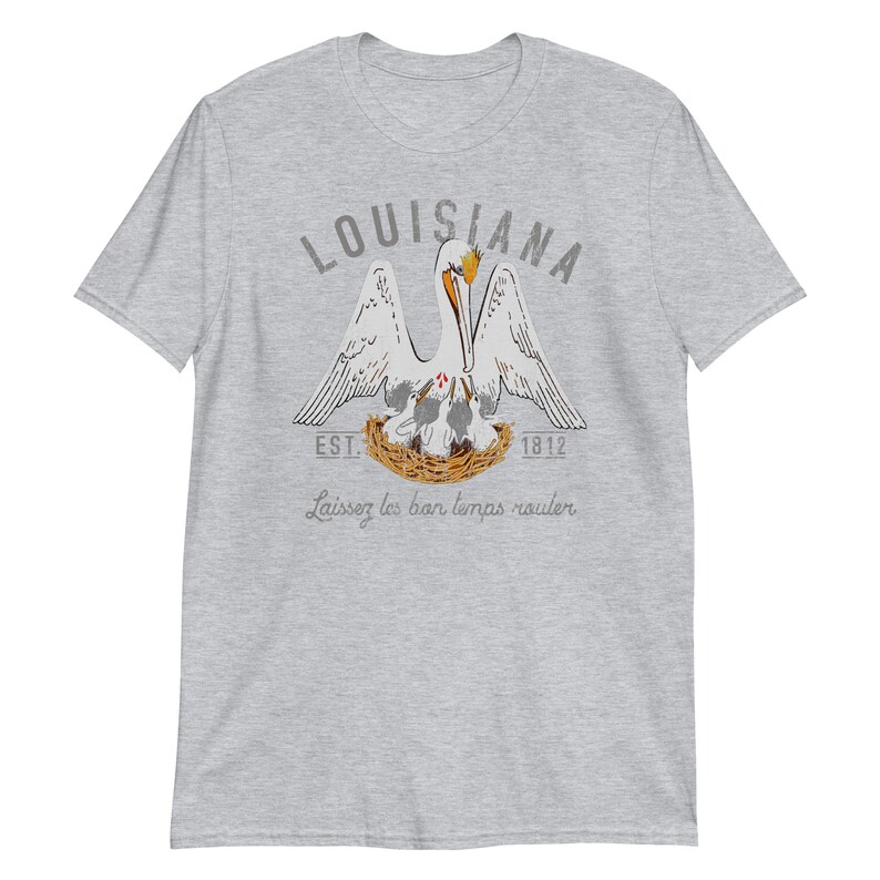 May include: A heather gray short-sleeve t-shirt featuring a graphic of a pelican in a nest with chicks. The text reads LOUISIANA, EST. 1812, and Laissez les bon temps rouler. The pelican is white with orange accents, and the nest is brown.