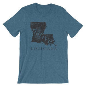 May include: A heathered blue short-sleeve t-shirt featuring a dark gray graphic of the state of Louisiana. The word native is printed in lowercase letters across the state shape, with the word LOUISIANA printed in capital letters below it.