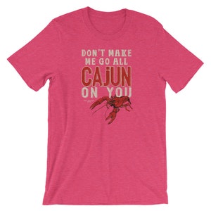 May include: A heathered pink short-sleeve t-shirt featuring a red crawfish graphic and the text "DON'T MAKE ME GO ALL CAJUN ON YOU" printed in a distressed white and red font. The shirt has a crew neckline and a standard fit design.