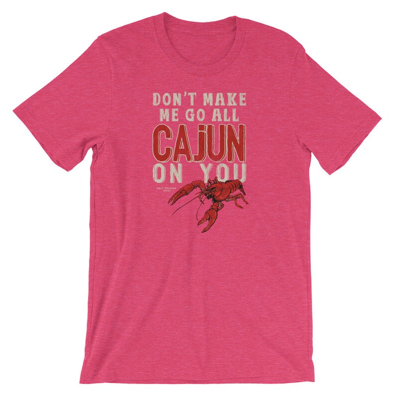 May include: A heathered pink short-sleeve t-shirt featuring a red crawfish graphic and the text "DON'T MAKE ME GO ALL CAJUN ON YOU" printed in a distressed white and red font. The shirt has a crew neckline and a standard fit design.