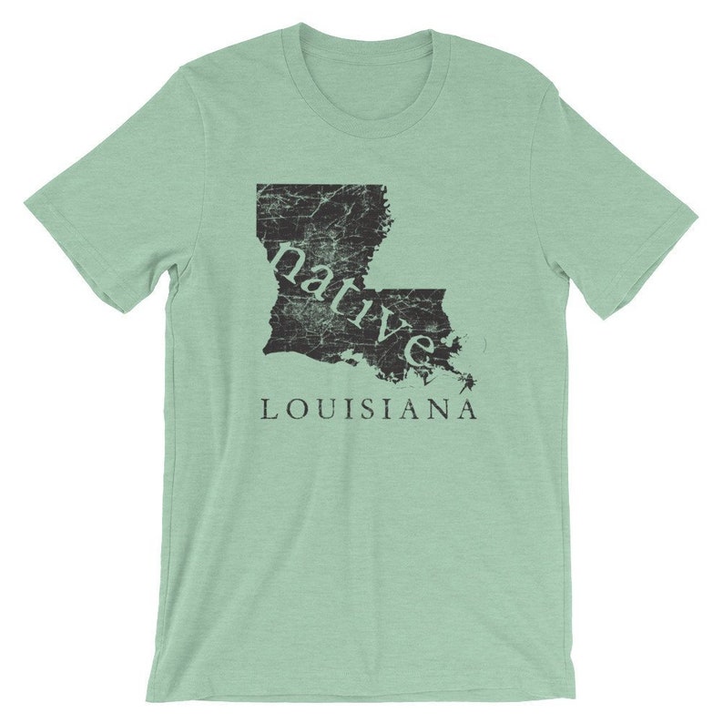 May include: A light green short-sleeve t-shirt featuring a black distressed graphic of the state of Louisiana. The word native is printed in a serif font across the map, and the word LOUISIANA is printed in capital letters below the map.
