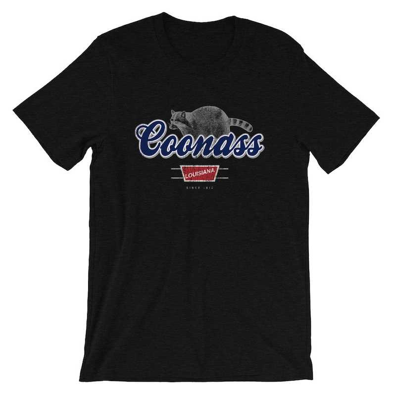 May include: Black t-shirt with a blue and white graphic of a raccoon with the text "Coonass" and "Louisiana" in a red rectangle with the text "Since 1812" underneath.