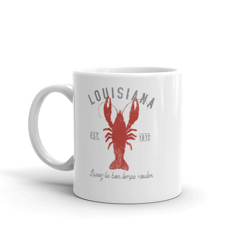 May include: A white ceramic mug featuring a red crawfish graphic in the center. Above the crawfish is the text "LOUISIANA" in gray, with "EST. 1812" on either side. Below the crawfish, the text "Laissez les bon temps rouler" is printed.