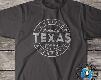 May include: A charcoal gray short-sleeve t-shirt featuring a circular white distressed graphic. The text reads: CERTIFIED PRODUCT OF TEXAS SINCE 1845 LOCAL-MADE AUTHENTIC. A small circular logo with a pelican illustration is in the bottom right corner.