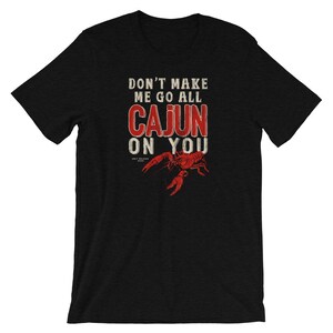 May include: A black short-sleeved t-shirt featuring a red crawfish graphic and the white and red text that reads: DON'T MAKE ME GO ALL CAJUN ON YOU. The design includes a small logo below the text that says UGLY PELICAN BRAND in white lettering.